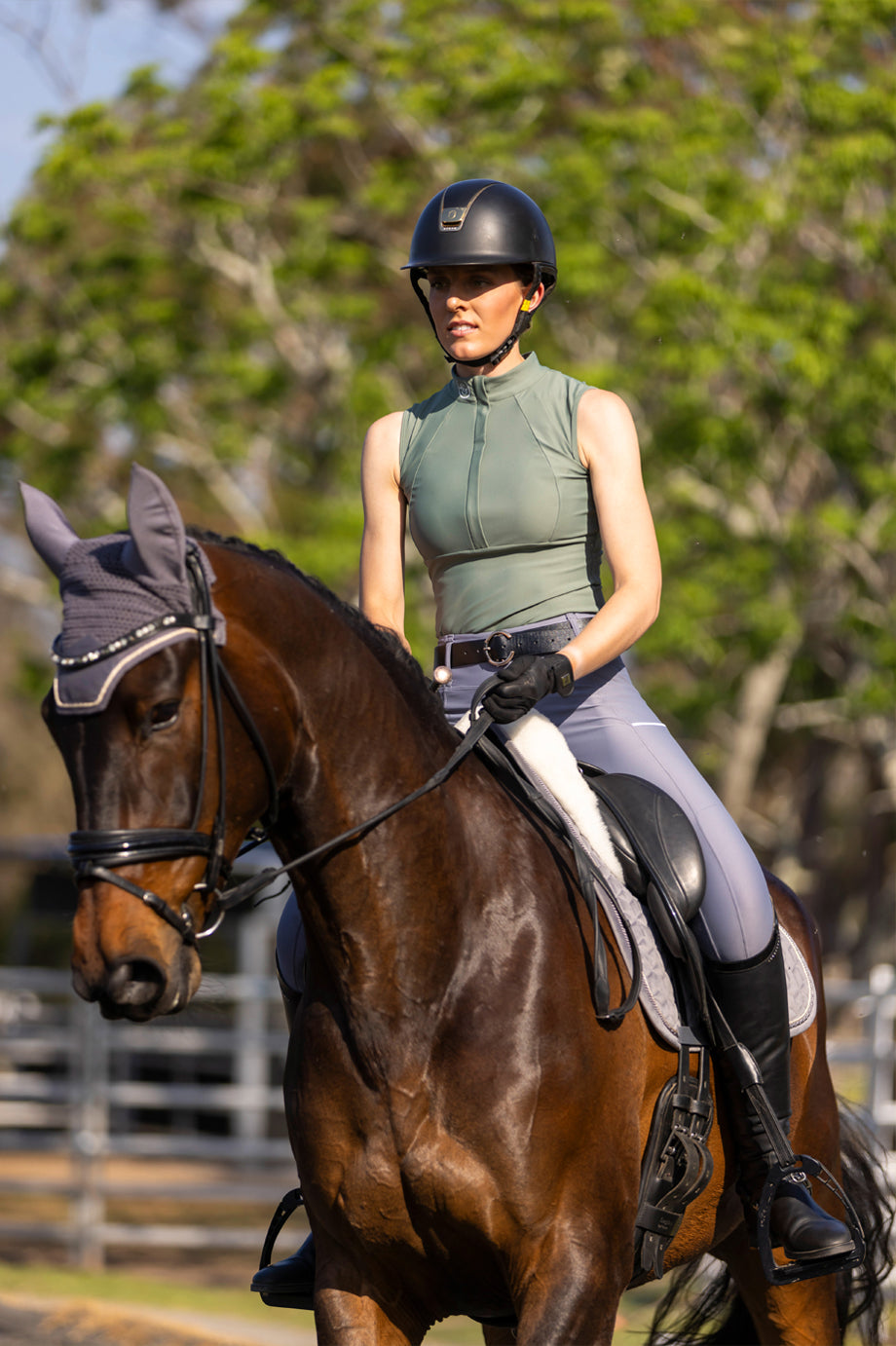 An image of a girl wearing the Sabi Equestrian Sleeveless Paradigm Design Base Layer in Viridescent Sage and the Sculpt Performance Hybrid Breeches in Silver Eclipse riding a brown horse